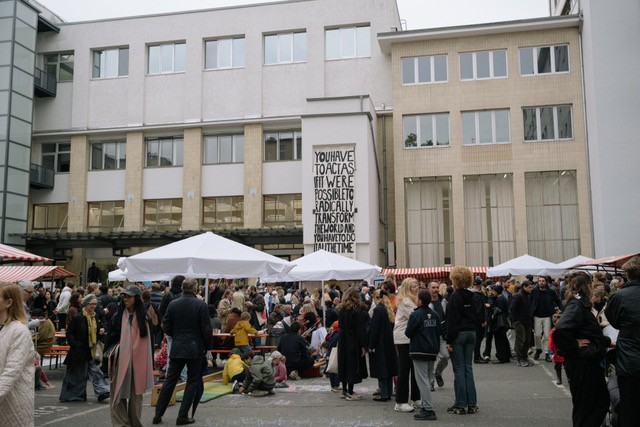 Viele Menschen stehen zwischen Ständen mit weißen und rot-weiß-gestreiften Überdachungen. Um sie herum sieht man verschiedene Häuser in den Himmel ragen.